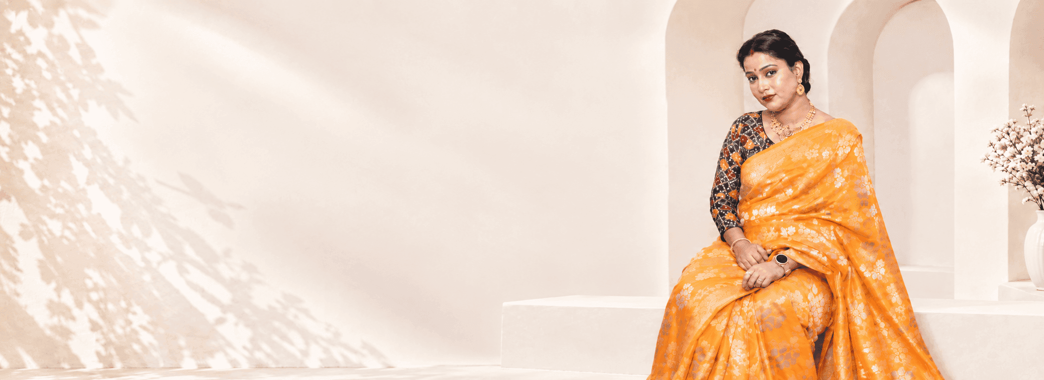 Model seated in a marigold Banarasi saree with silver floral zari, in a sunlit arched alcove
